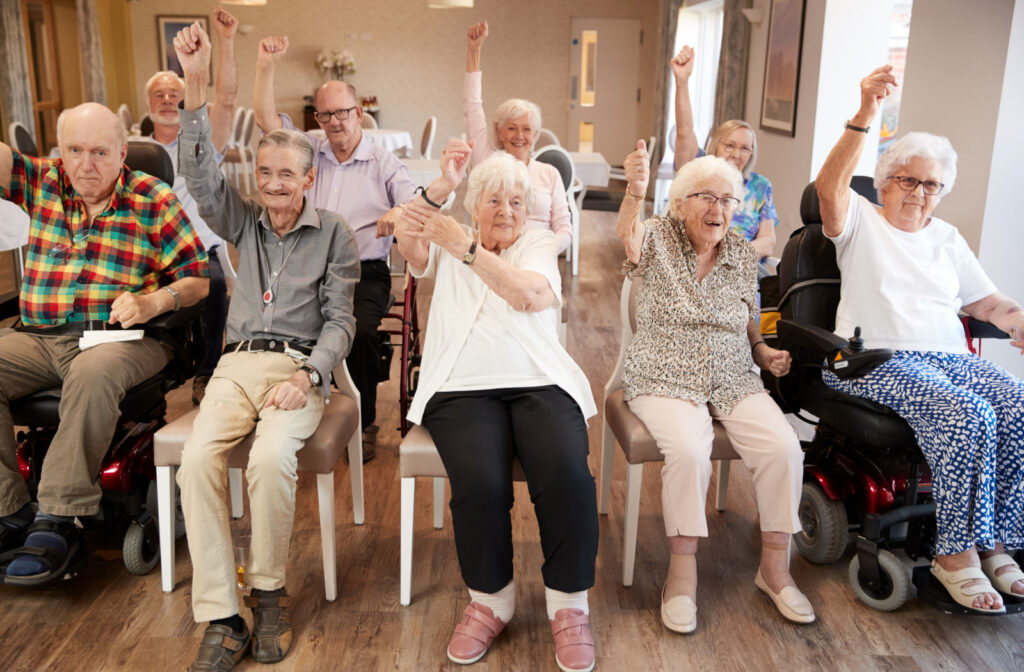 A group of seniors sitting in chairs, doing seated exercises with one arm raised in the air.