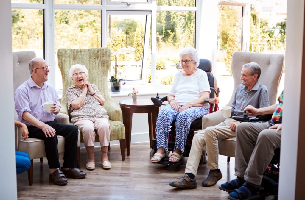 A group of older adults drinking tea and laughing, sitting in a semicircle in armchairs within an assisted living facility.
