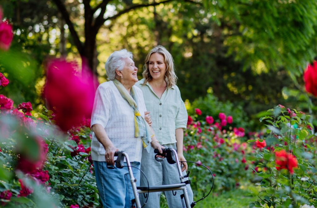 Caregiver supporting senior resident when taking her for walk with walker in park in summer.