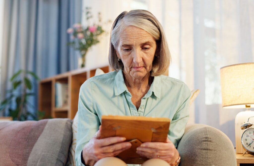 A senior sitting on their living room couch holds a framed photograph in their hands, looking at it forlornly