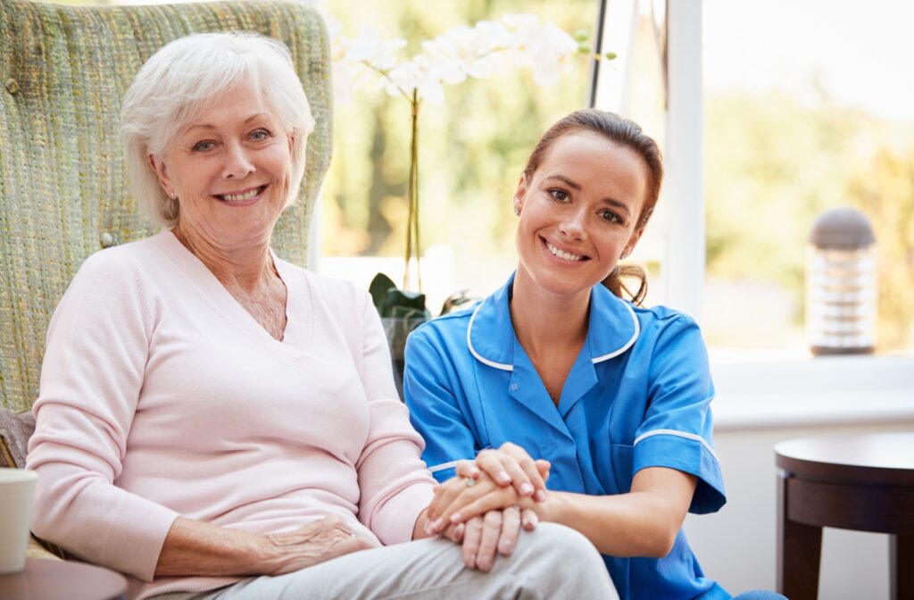 An older adult and a caregiver holding hands and smiling at the camera in a common area of their assisted living community.