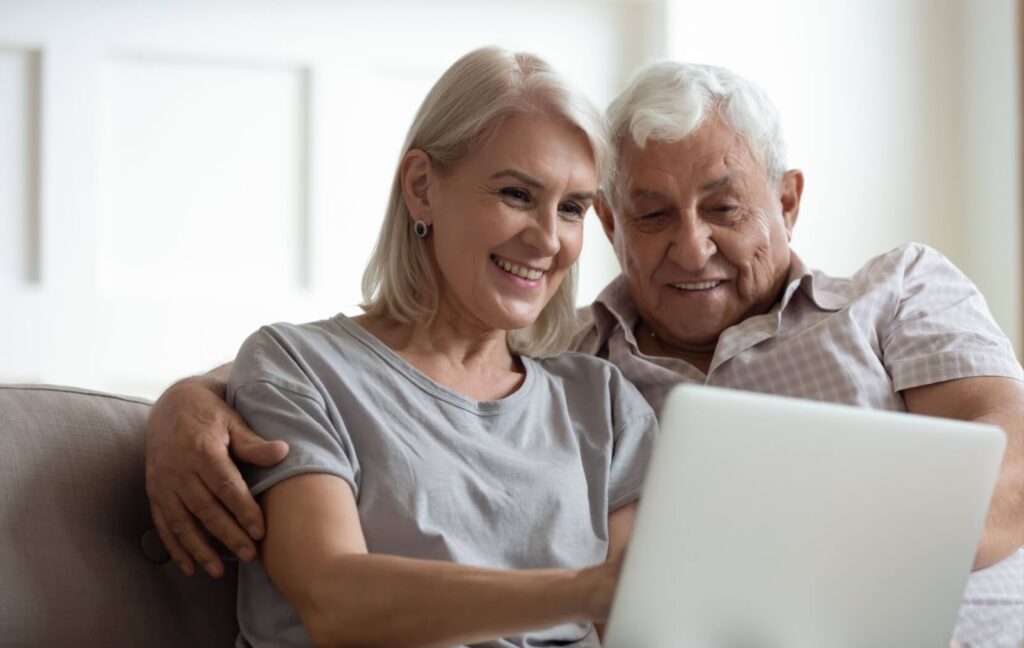 A couple of older adults sit on a couch while they use a laptop to video call their adult children who live in a different location.