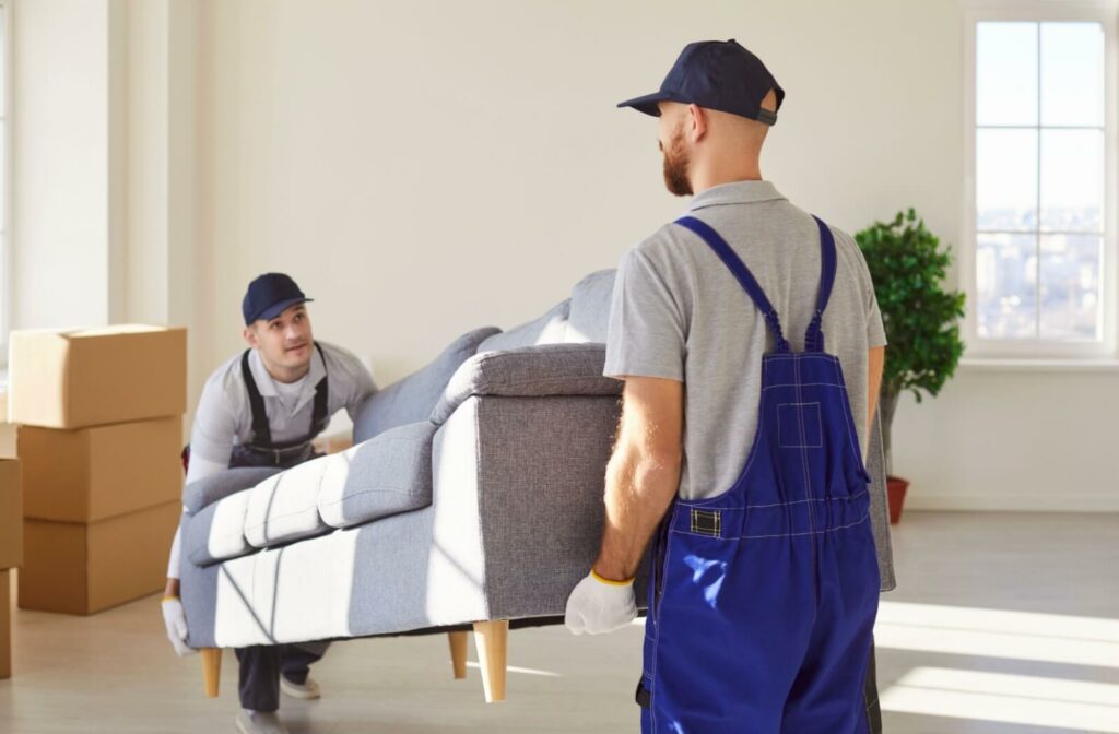 Two professional movers in overalls lift a couch, prepping to load it into a truck to assist in moving a client’s belongings