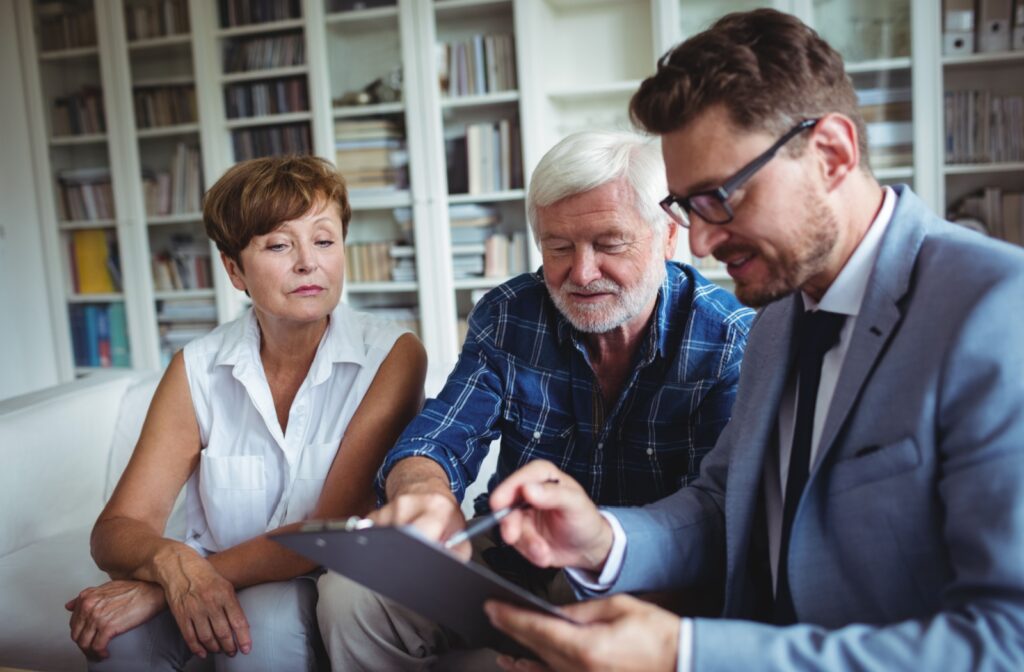 An adult child and their senior parent meet with an assisted living intake coordinator while sitting on a white couch in front of a wall of bookshelves.