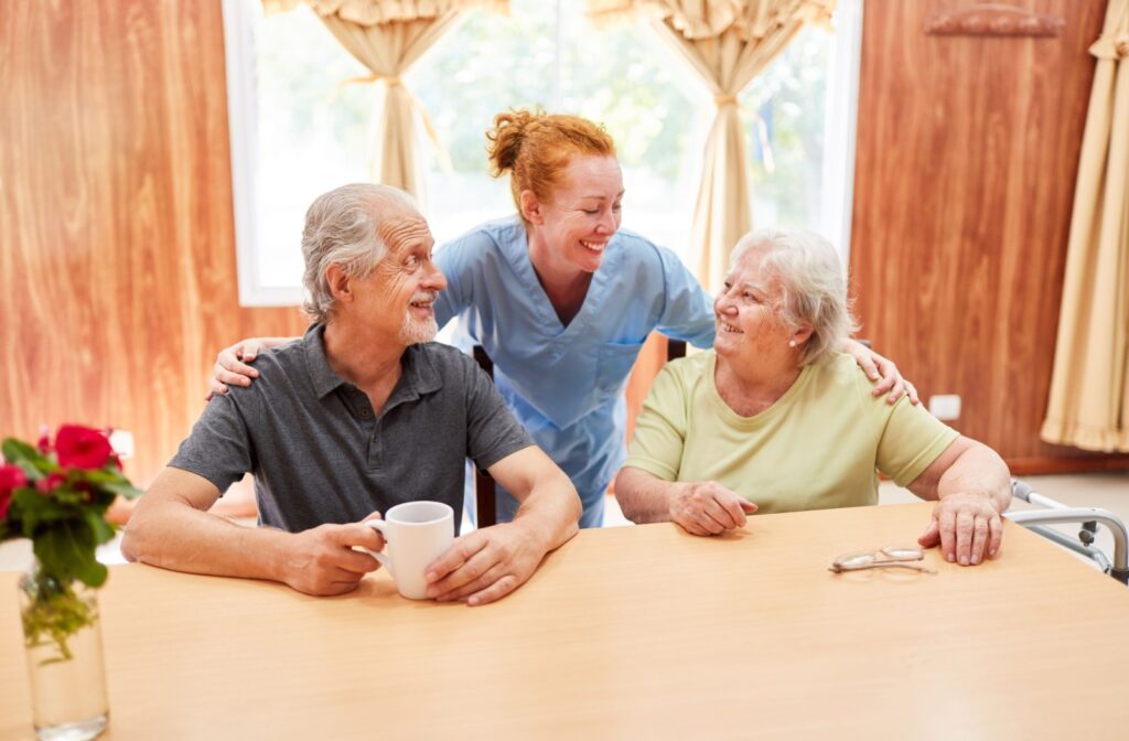 A caregiver checks in on an older couple at a breakfast table while standing behind them in assisted living