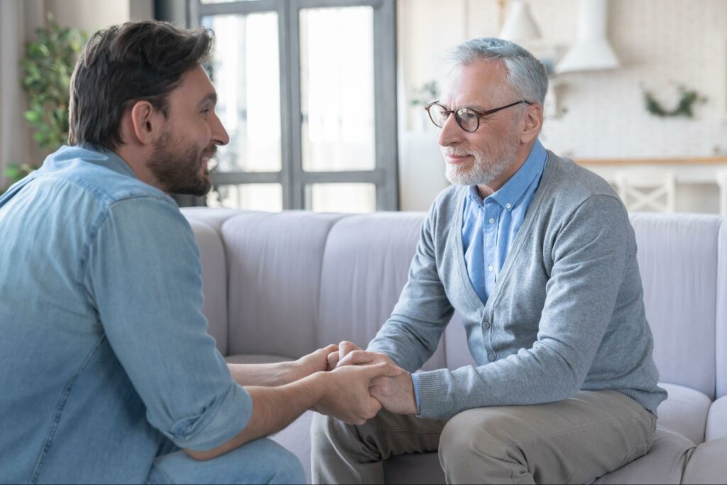 An adult child and their senior parent sit across from each other, holding hands to offer comfort during an important conversation.